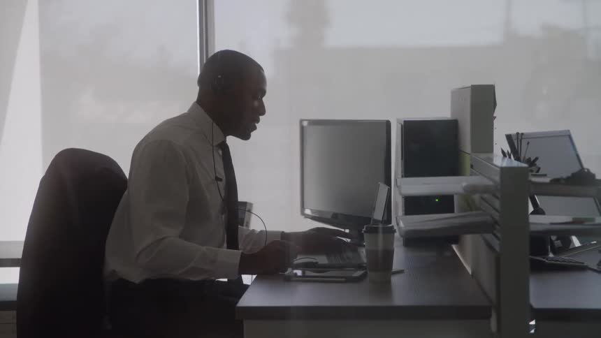 The video is a silhouette of an african american man in a white shirt and tie sitting at a desk and working on a computer in a brightly lit office he is wearing a headset suggesting he is on a call or using it for communication while focusing on his scr