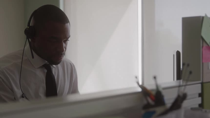 A young man wearing headphones types on a computer while sitting behind a desk in an office at midday