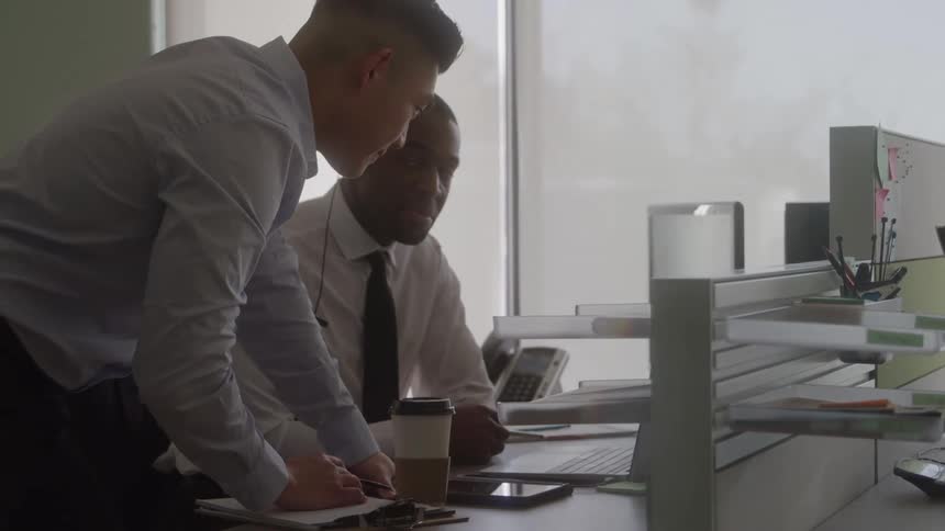 The video is an overtheshoulder shot of two men in an office cubicle area one man wearing a headset is seated at a computer while another man stands over him pointing at a document on the desk