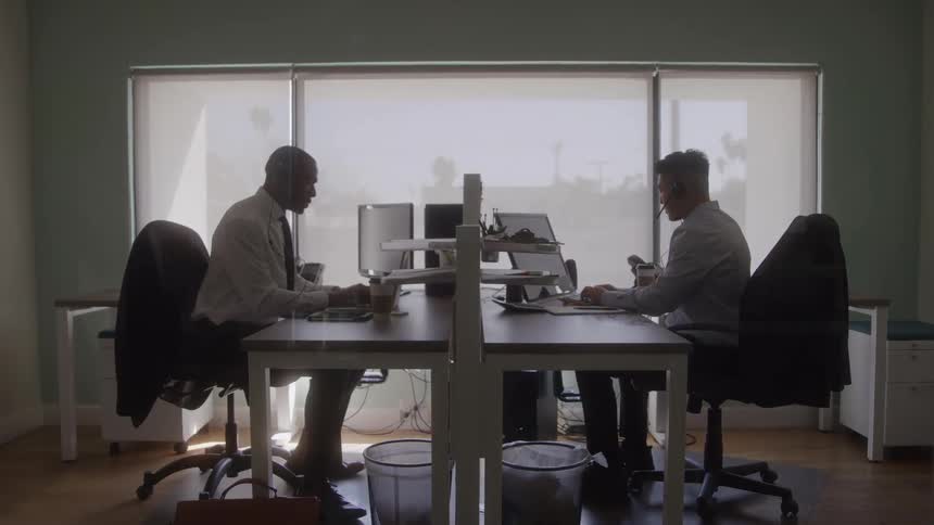 Two men are sitting at desks facing each other in an office