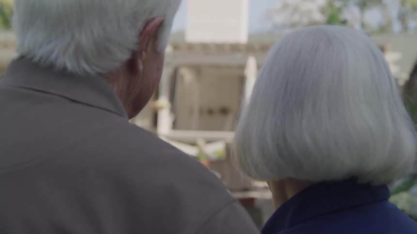 An elderly couple stands on a driveway looking at a suburban home smiling at each other happily at midday