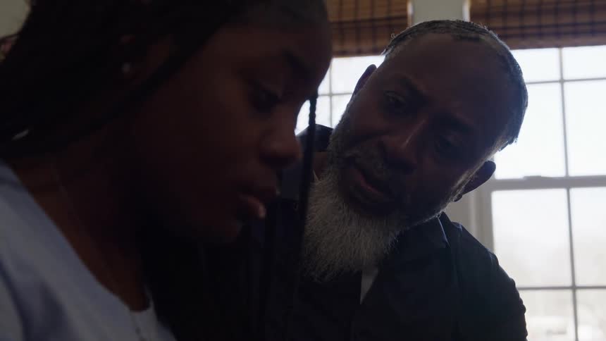 A black man with a gray beard leans in to speak gently with a sad looking black girl by a bright window