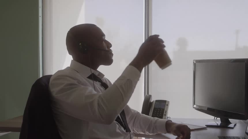 The video shows an office worker a man in a white shirt and tie sitting in silhouette at his desk in front of a bright window he is wearing a headset and lifts a travel mug to drink suggesting he is busy working in a call center or open office environ