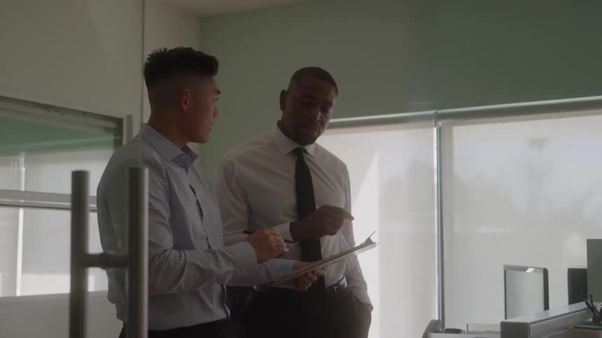 Two men stand inside an office wearing longsleeve office shirts having a discussion with one holding a folder at midday