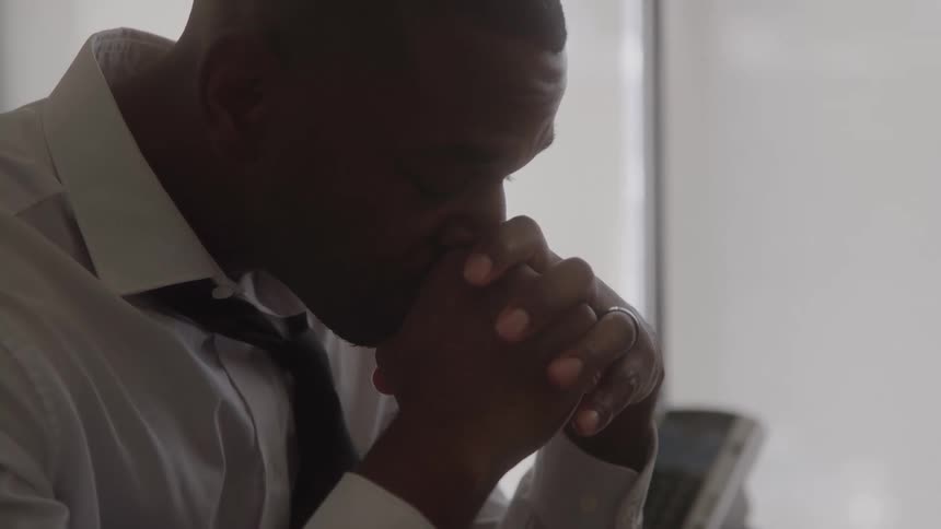 A stressed man sits with his head bowed and hands clasped at his desk in an office
