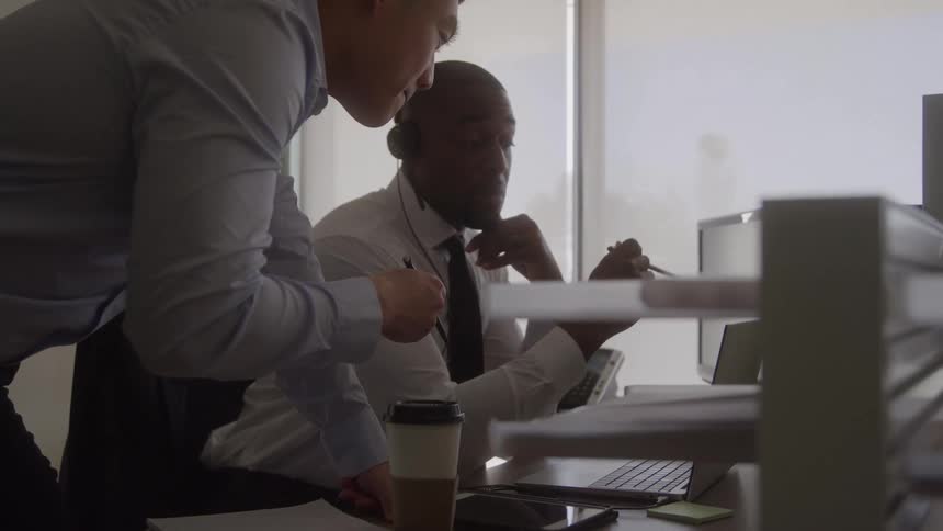 The video is a medium closeup of two office workers one african american and one asian standing and reviewing something on a computer screen at a desk the african american man is seated and wearing a headset while the asian man leans in with a pen a