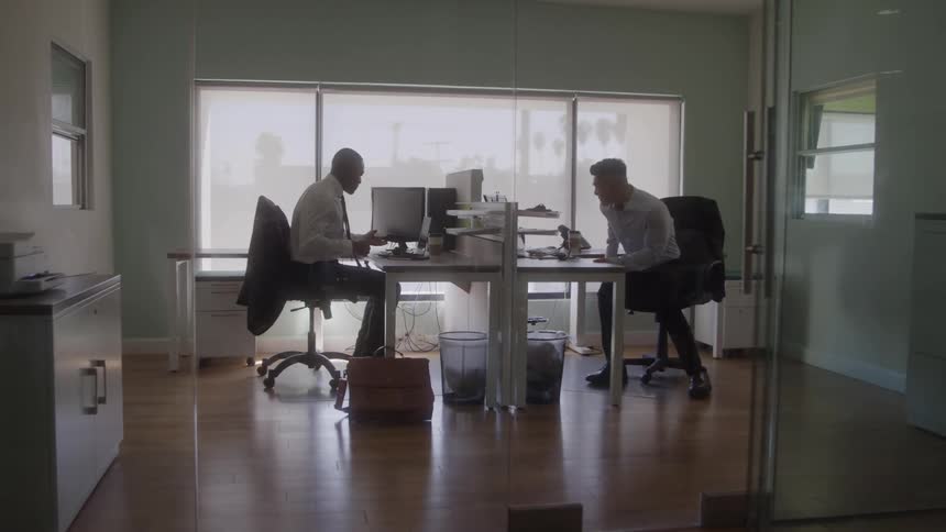Two individuals seated across from each other at office desk with computers papers trash bins and side cabinet