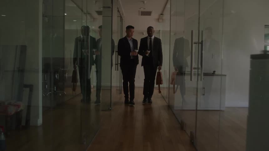 Two men carrying disposable coffee cups walk down a hallway lined with glass doors