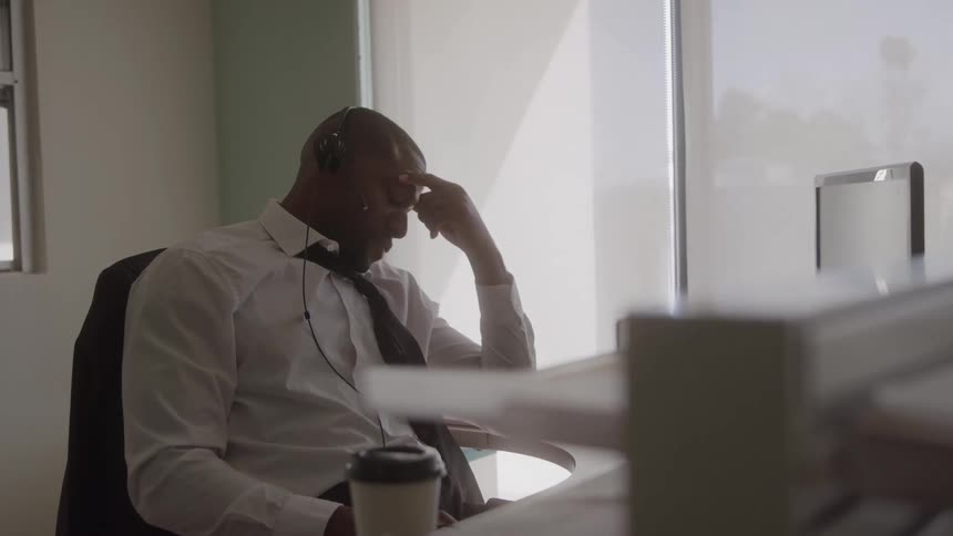 A stressed office worker with a headset pinches the bridge of his nose while sitting at his desk