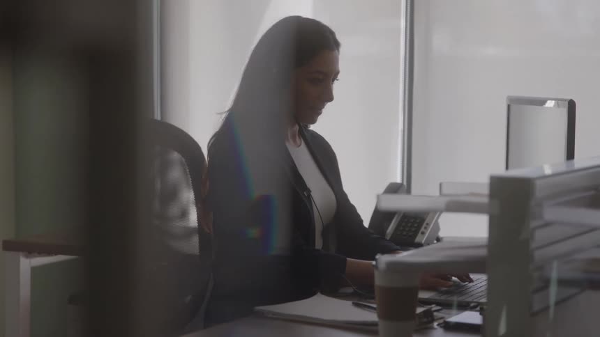 The video is a medium closeup of a woman with dark hair sitting at an office desk looking down and working on a computer or telephone the scene is brightly lit by large windows in the background which silhouettes her figure and creates a professional