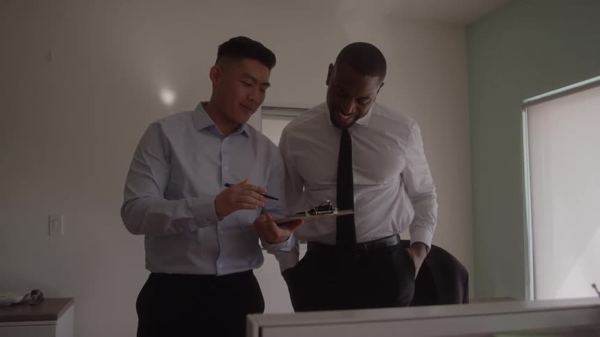 In an office a man is holding a pen and a clipboard talking and showing the clipboard to another man