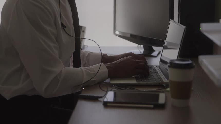 A man in a shirt and tie with an earpiece types on a laptop at his desk with a coffee and smartphone nearby