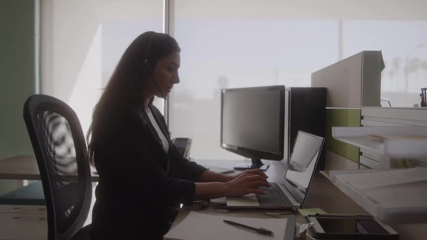 The video is a medium shot of a professional woman with dark hair in a black blazer and headset sitting at a desk and typing on a laptop she appears focused and busy working in a brightly lit modern office with a monitor and a bright window in the back