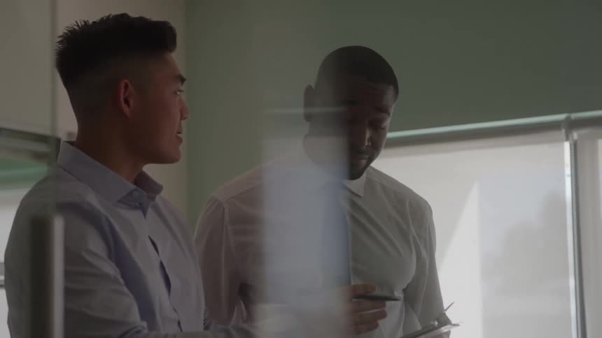 The video shows a medium closeup of two men one asian and one african american in business attire standing close together and engaged in a serious conversation or discussion at an office the shot is framed to look through a glass partition with the