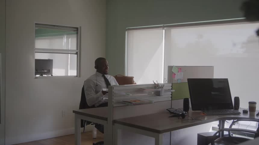 The video is an interior office shot where a man in a lightcolored shirt is walking past a colleague who is seated at a desk the seated african american man turns to smile while talking on a headset at his cubicle which is brightly lit by the backgroun