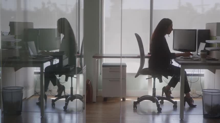 The video is an interior shot of a woman in an office setting sitting at a desk and working on a computer the scene is brightly lit by large windows with blinds and the womans figure is reflected in the glass partition of the office creating a double