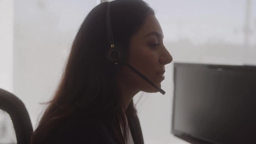 A woman wearing a headset speaks while working at her computer in an office