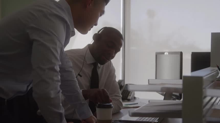 In an office a man is sitting with a pen in his hand talking to another man who is standing beside him