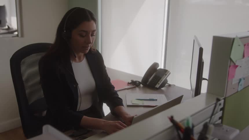 Woman wearing a headset works on a laptop inside a bright office