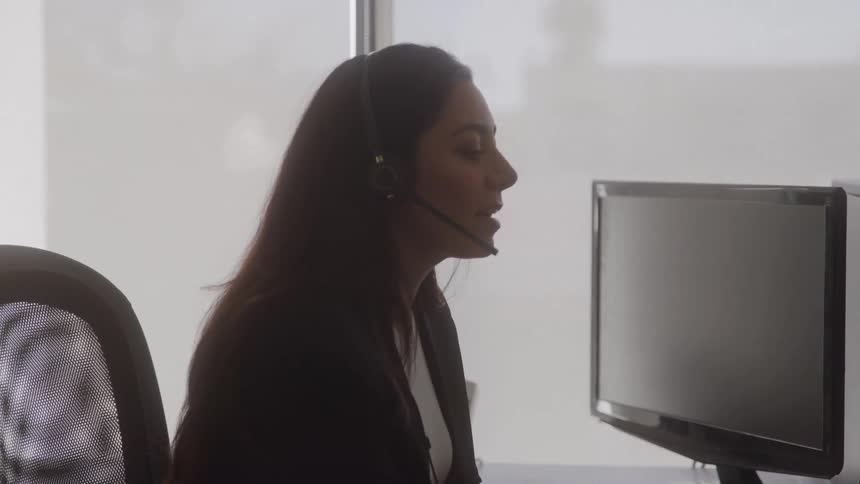 The video shows a medium closeup of a woman in a dark blazer wearing a headset as she works at an office desk she is looking directly at a computer monitor indicating she is likely engaged in an online meeting or a customer service call