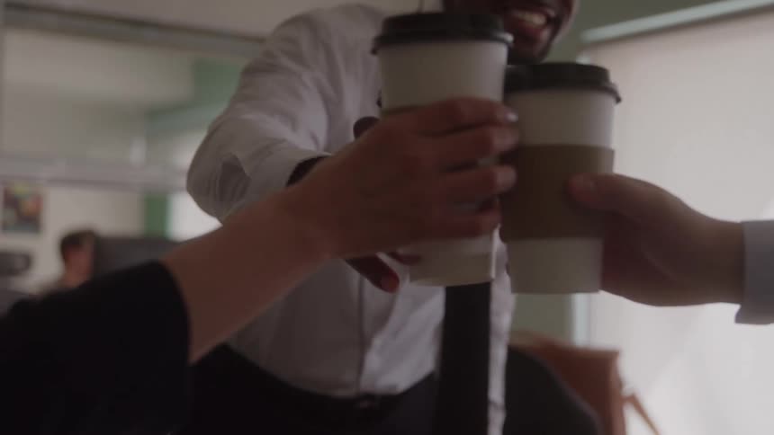 The video is a medium closeup of a smiling african american man in a white shirt and tie wearing a headset in an office cubicle the scene cuts to a clinking of coffee cups in an office setting as a sign of a celebratory or friendly toast
