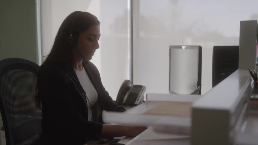 The video is a medium shot of a woman with dark hair and a headset working at a desk in an office she is seen typing on a laptop with a secondary monitor visible next to her likely working in a customer service or administrative role