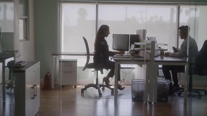 The video is a wide shot of two people working at desks in a modern brightly lit office with large windows covered by light blinds the figures are mostly silhouetted against the bright background with the woman sitting prominently in the foreground wor