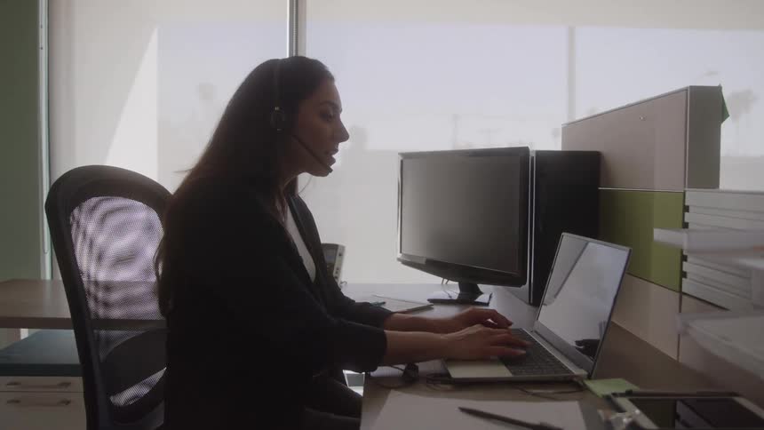 Woman sitting in her office cubicle types while talking on a headset