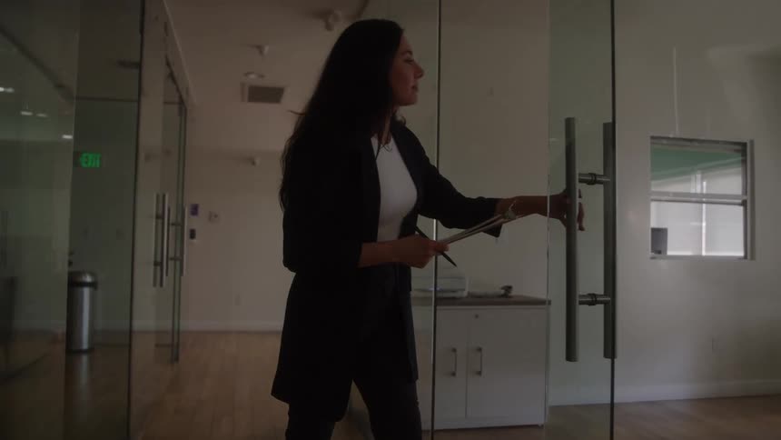 The video is an interior shot of a professional woman in a dark blazer walking down a hallway lined with glasswalled offices she stops at an office door which has a silver handle holds a clipboard and turns her body to enter revealing a