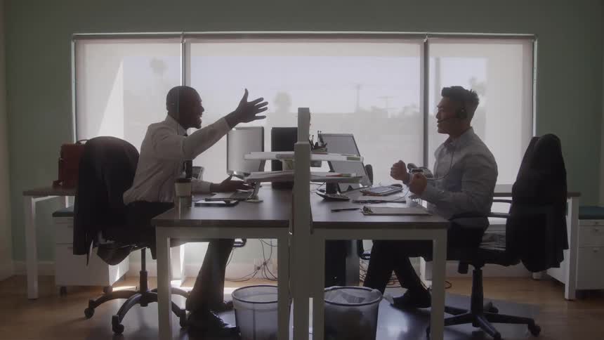 Two male coworkers share a fist bump across their desks in a bright office