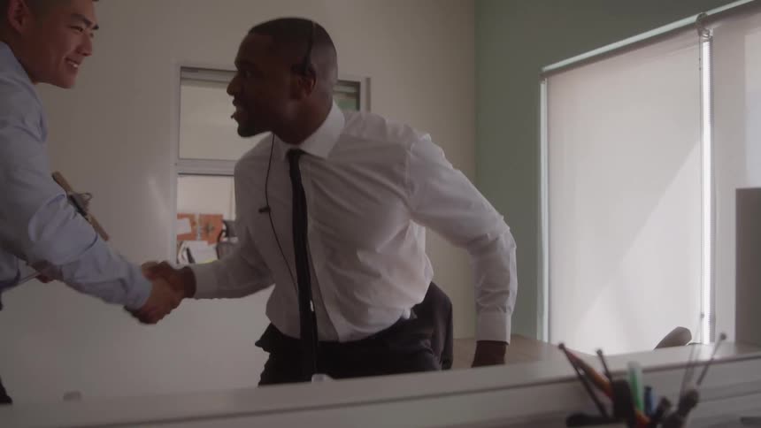 The video is an interior office shot where an african american man in a white shirt and tie wearing a headset is sitting and laughing heartily at his cubicle desk an asian colleague holding a clipboard then approaches and the two men rise to shake han
