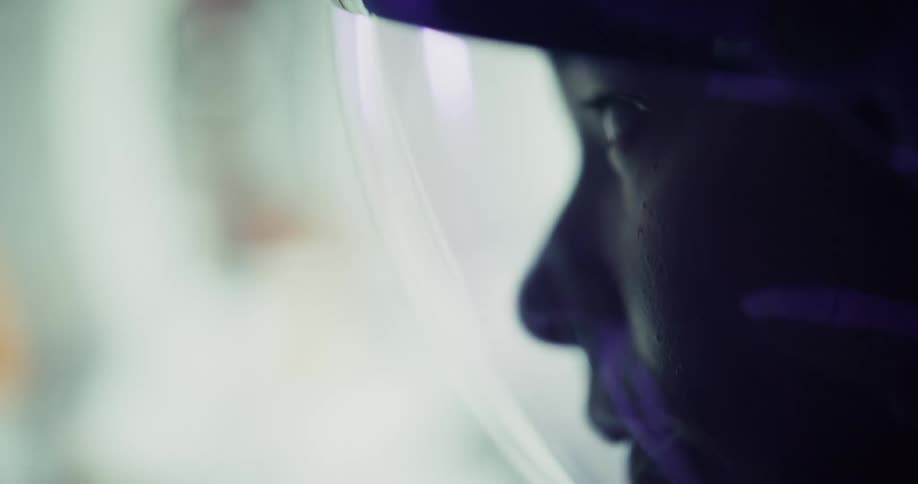A close profile view shows a womans face reflected in the clear visor of a dark helmet in a softly lit dramatic interior setting