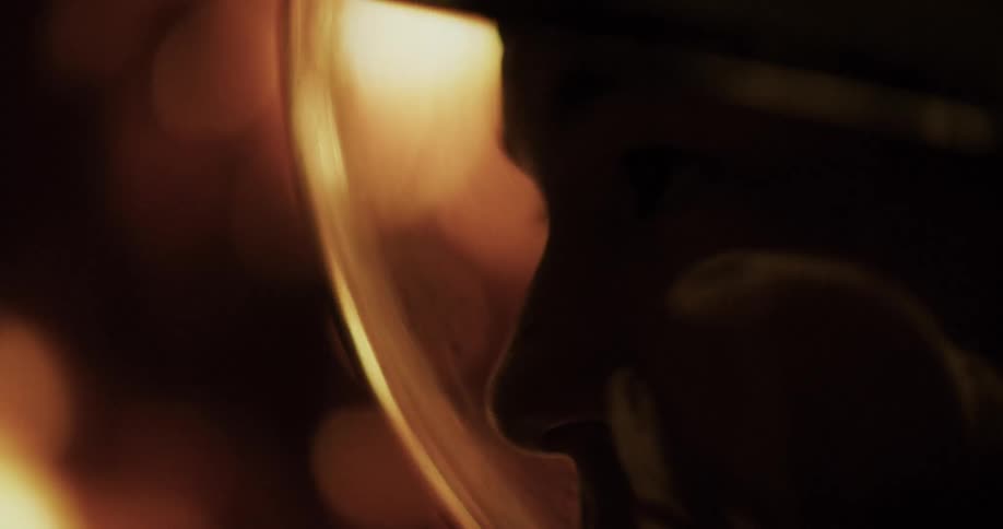 A close profile of a face in deep shadow with an orange light reflecting off the visor of a space helmet
