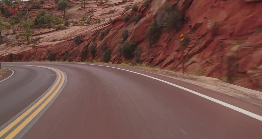 The video is a drivers perspective shot traveling along a winding reddishbrown paved road the road is flanked by steep red rock formations and scattered greenery under a bright cloudy sky