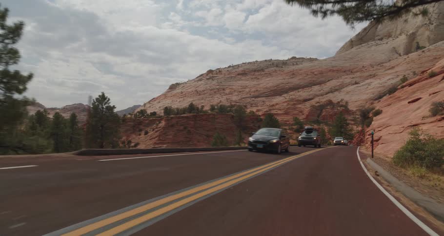 Vehicles travel along a winding road through rocky desert mountains under a cloudy sky