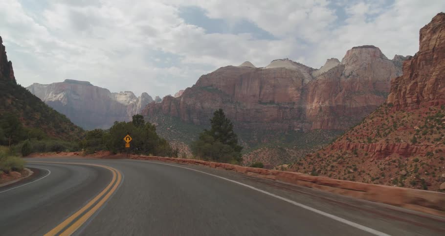 Winding road surrounded by trees and rocky mountains under a partly cloudy sky