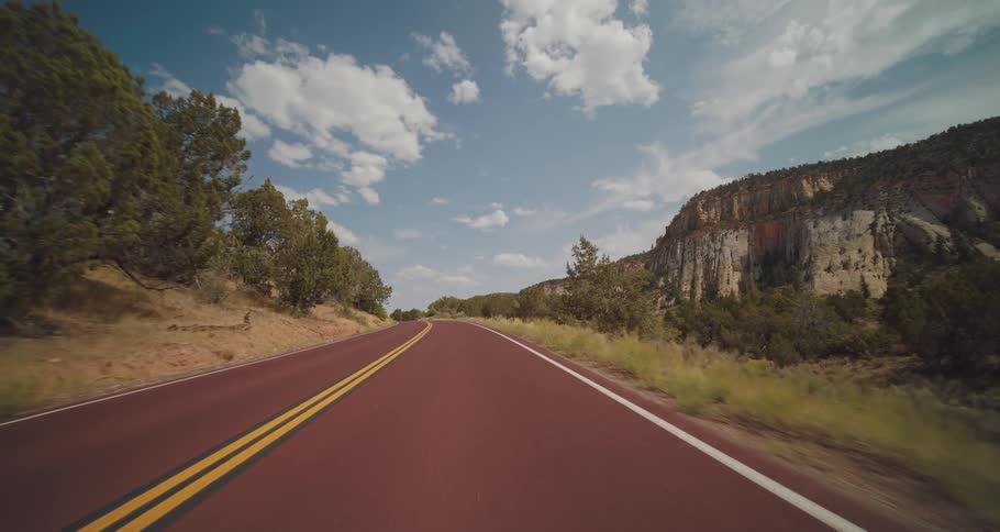 A vehicle view looking down a red asphalt road through a mountain canyon shows a car driving away surrounded by light colored rock cliffs green trees and a bright blue sky with white clouds