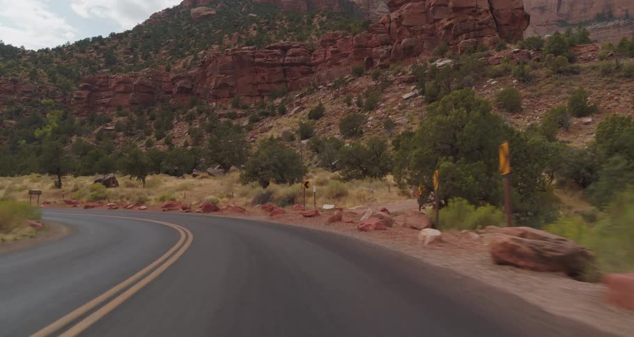 A winding road leads through rocky mountains and dry brush under a cloudy sky