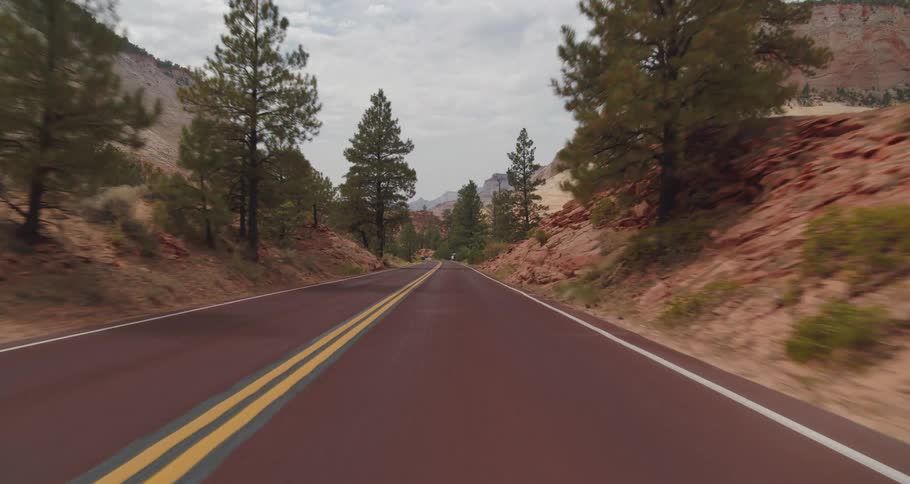 A vehicle view drives along a red asphalt road through a mountain canyon passing other cars and surrounded by towering tan and reddish rock cliffs and thick green trees under a bright overcast sky