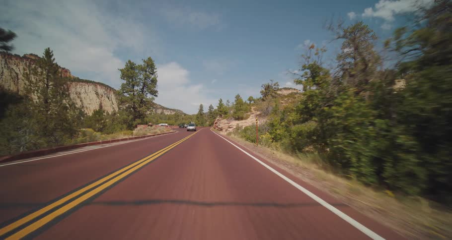 A scenic drive along a reddish road winding through a desert canyon flanked by rocky cliffs and pine trees