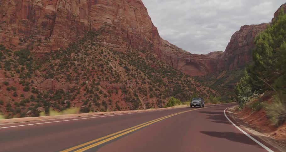 A vehicle view drives along a curving two lane road through a deep mountain canyon with towering red rock cliffs slopes covered in green brush and oncoming cars under an overcast sky