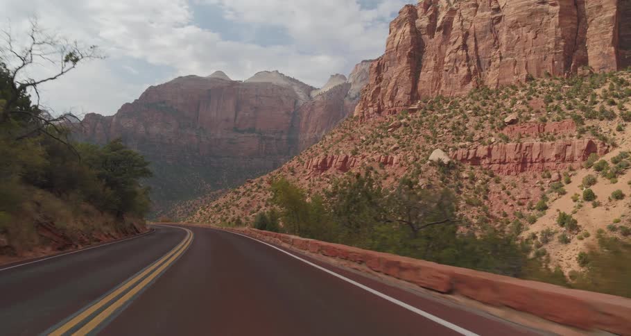 A vehicle view drives along a curving two lane road through a deep mountain canyon with towering red rock cliffs slopes covered in green brush and oncoming cars under an overcast sky