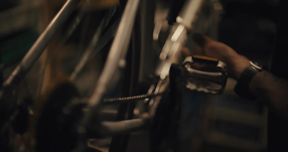 In a dimly lit workshop a persons hand spins the pedal of a bicycle causing the rear wheel and gear cassette to turn