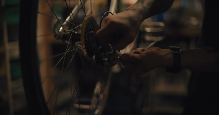 A close view shows the hands of a mechanic working on the rear hub and spokes of a bicycle wheel in a dark dimly lit repair shop