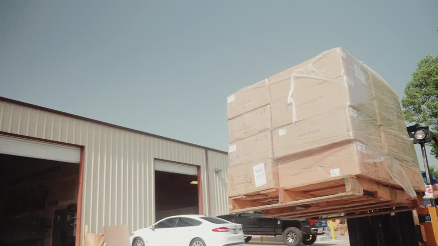 A forklift lifts a pallet stacked with boxes outside a warehouse at midday with two cars parked by the entrance
