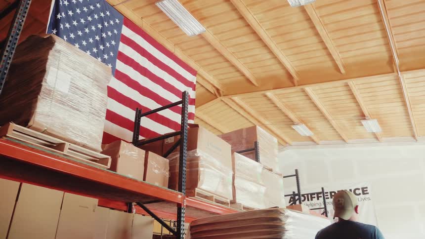 Worker arranges boxes in a warehouse with the united states flag in the background