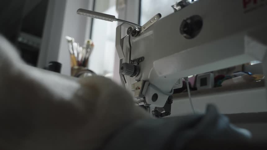 A close view of a white industrial sewing machine shows the needle and foot with fabric being stitched below under the bright focus of an attached light