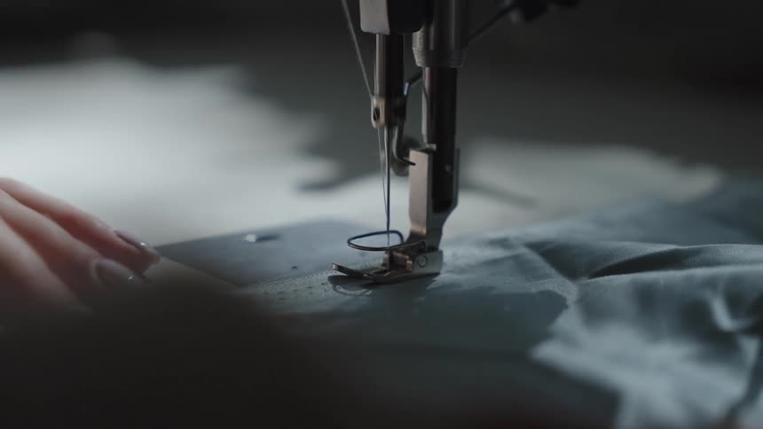 A close view shows the hands of a woman guiding blue fabric under the needle and presser foot of an industrial sewing machine as it stitches a seam