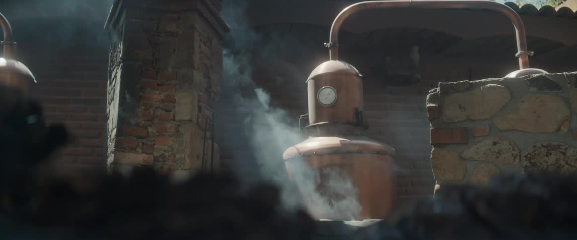 A large copper distillation still hangs suspended steam billows around the apparatus the still is connected to curved piping two stone and brick structures frame the scene