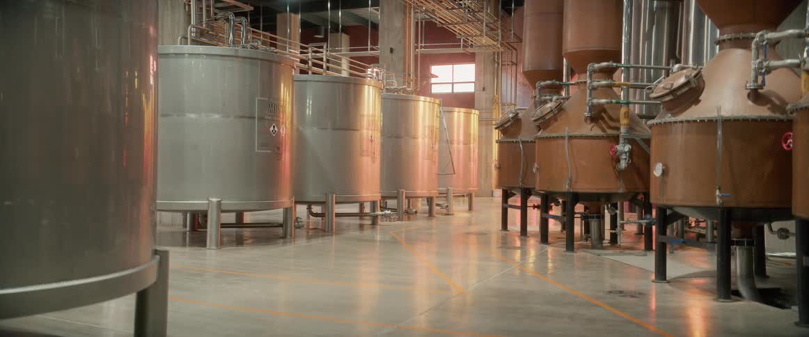 A view inside a clean industrial building several rows of tall silver metal tanks stand on the left multiple pieces of large brown copper processing machinery fill the right utility pipes run across the ceiling